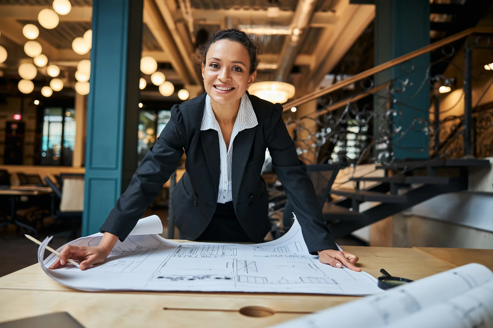 Lächelnde Architektin mit Bauplänen am Tisch als Beispiel für eine professionelle Website
