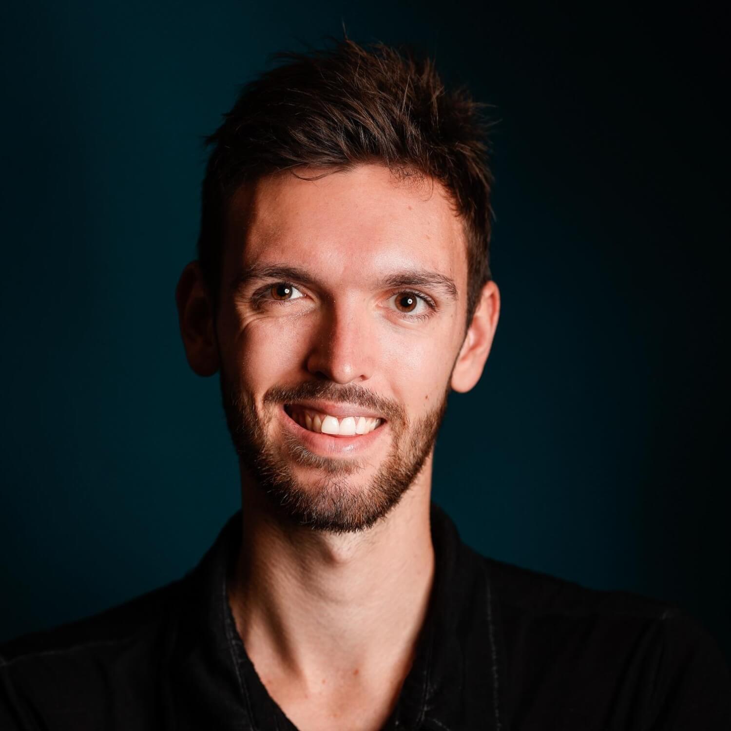 Nahaufnahme eines lächelnden Mannes mit Bart vor dunkelblauem Hintergrund, Studio-Porträt mit weichem Licht.