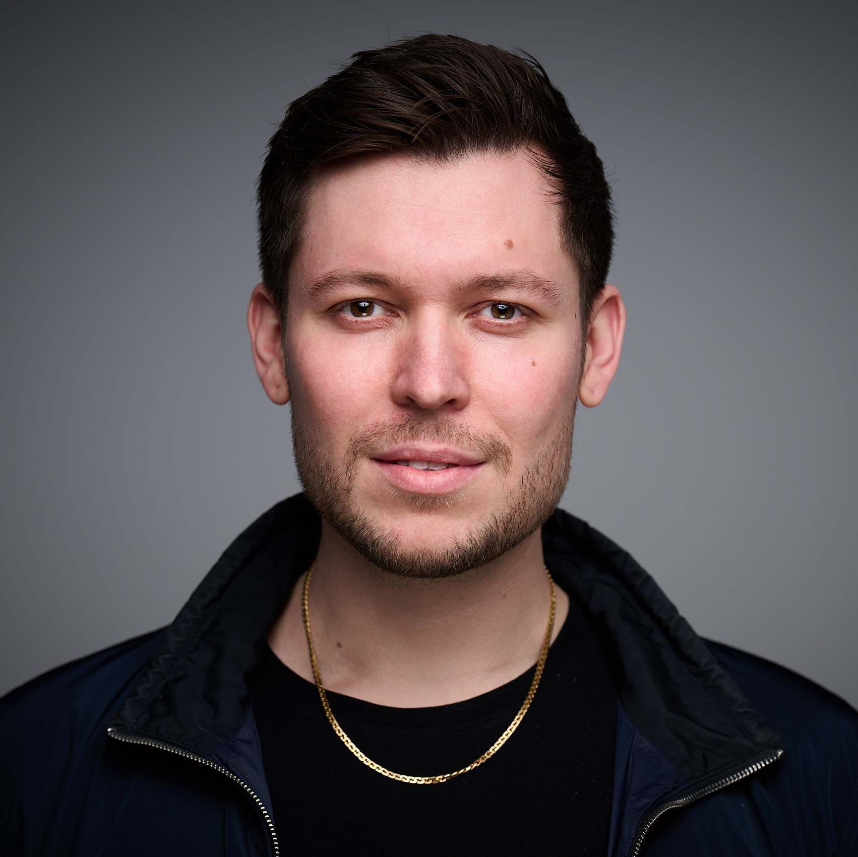 Nahaufnahme eines Mannes mit dunklen Haaren, leichtem Bart und goldener Kette vor neutral grauem Hintergrund.